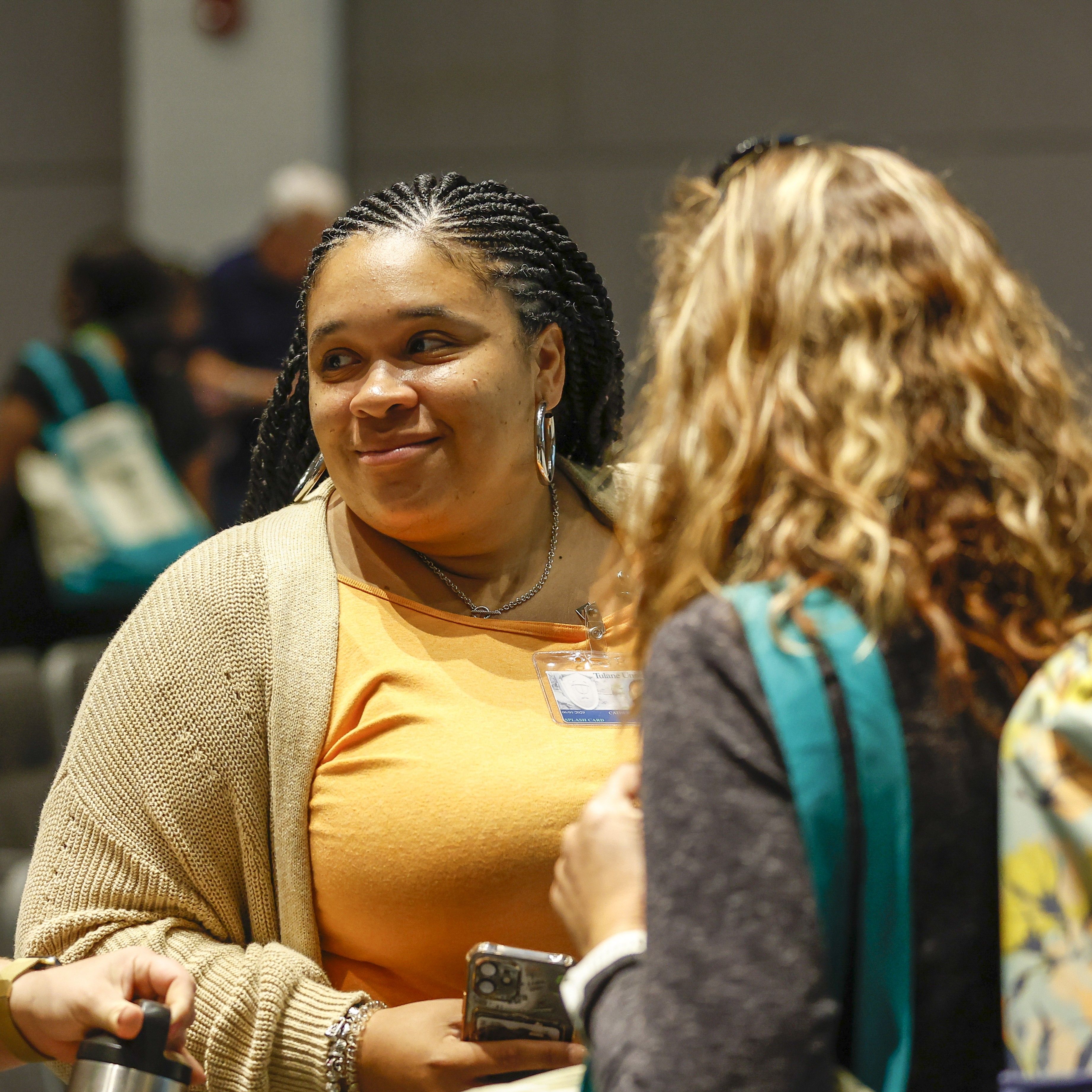 Smiling woman with braided hair and an orange shirt talks to another woman.
