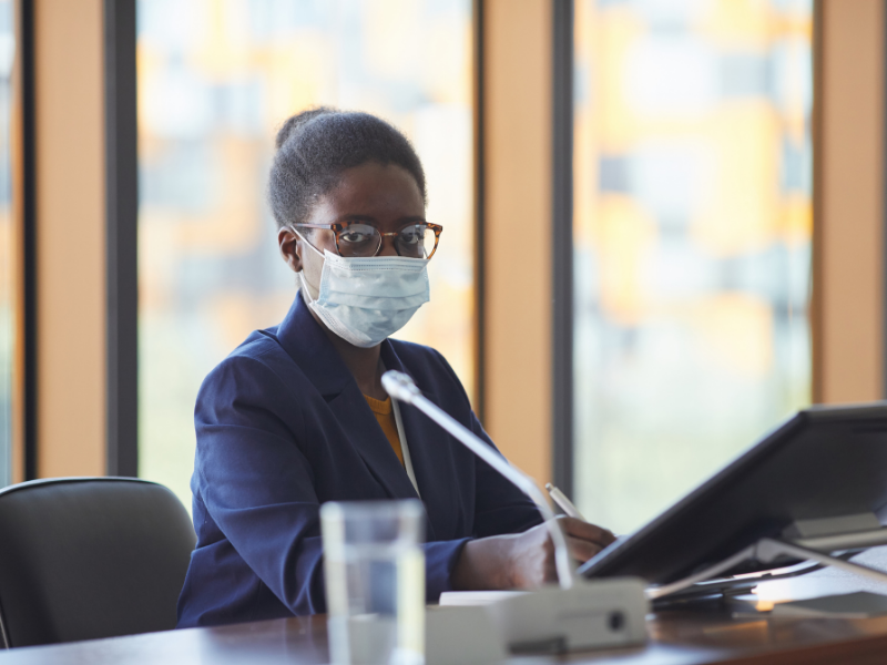 Stock photo of an African-American woman wearing a mask and seated at a table presenting from a laptop.