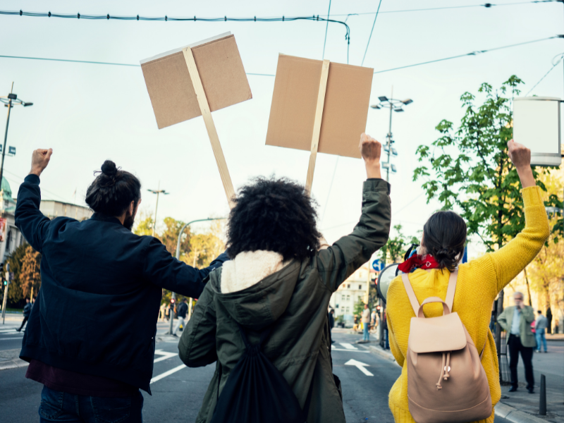 Three diverse individuals participate in a street protest with their backs to the camera, carrying signs, and raising their fists in the air.